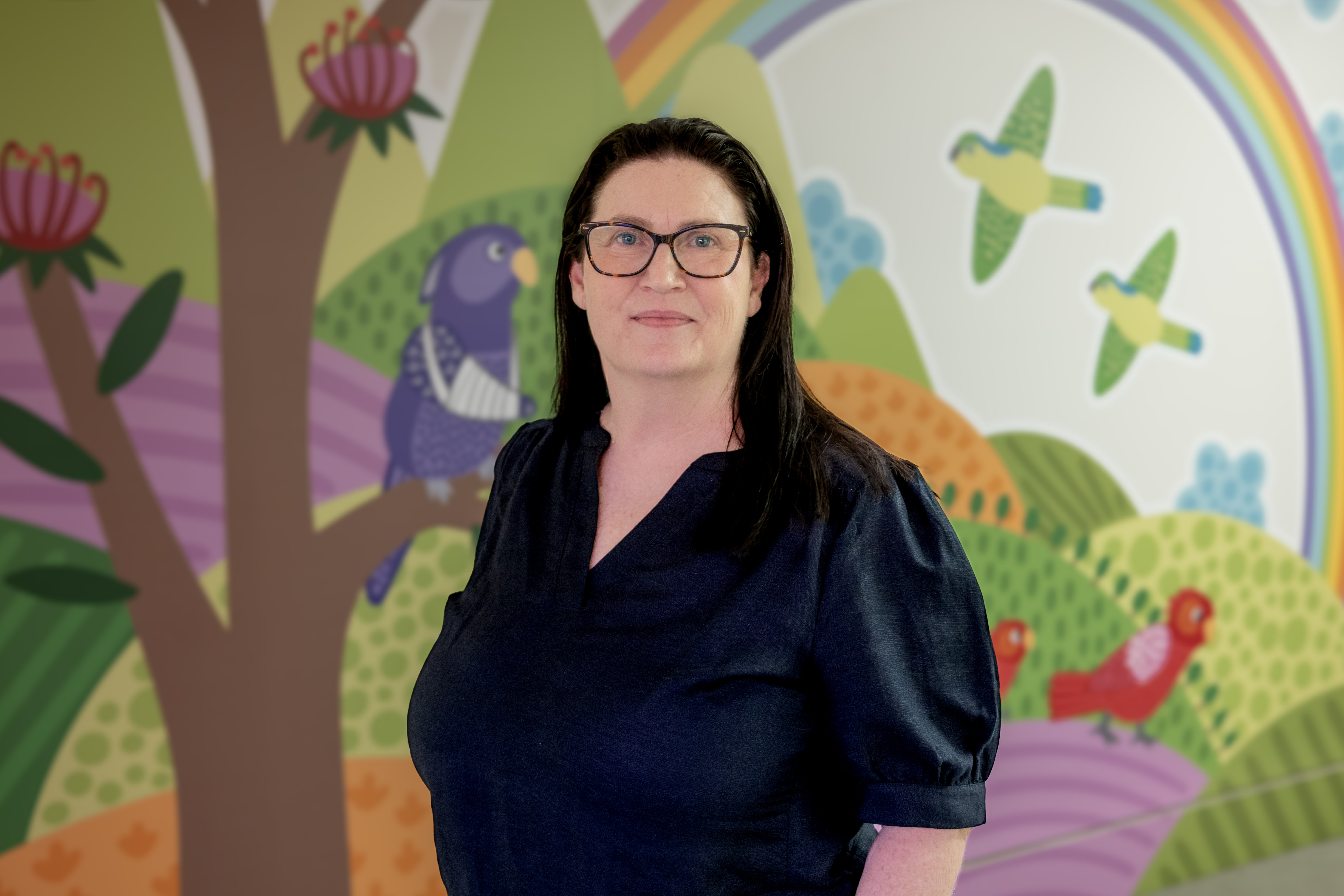 A woman with dark hair and black rimmed glass standing in front of a bright patterned wall