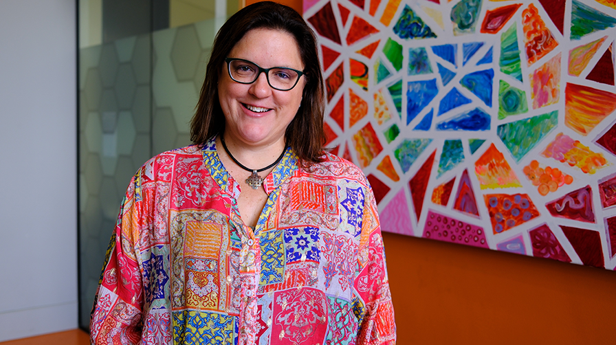 A woman with dark hair and black rimmed glass standing in front of a bright patterned wall