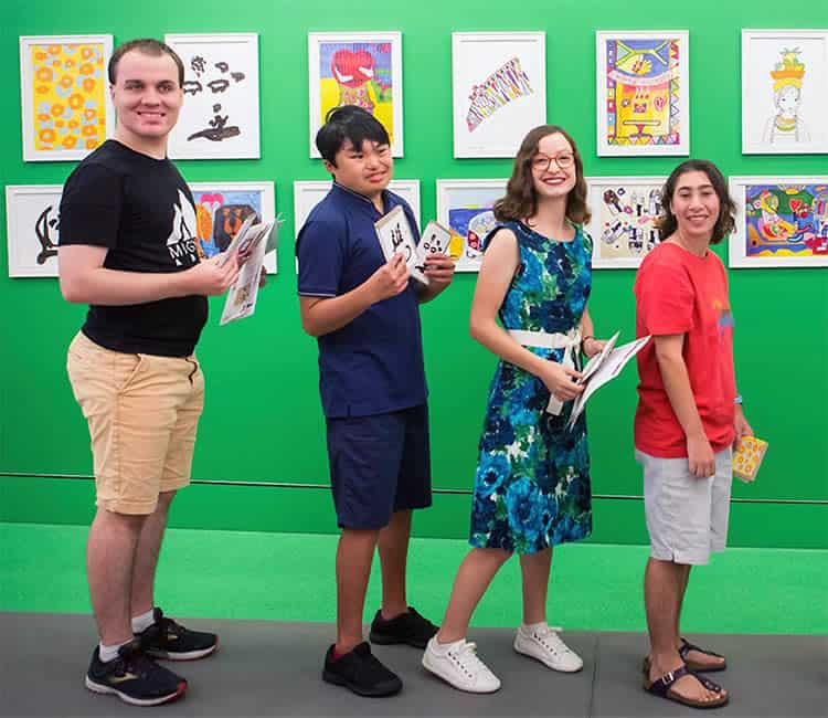 Four young people holding artwork and standing in a line in front of a green wall with artwork hanging on it