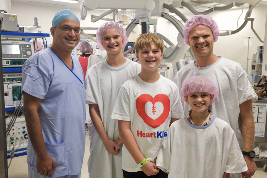 Smiling group of people (2 men, 2 high school aged children and one primary aged boy wearing scrubs standing in an operating theatre.