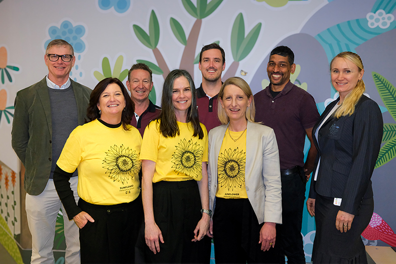 A group of 8 people standing in front a decorative wall. 